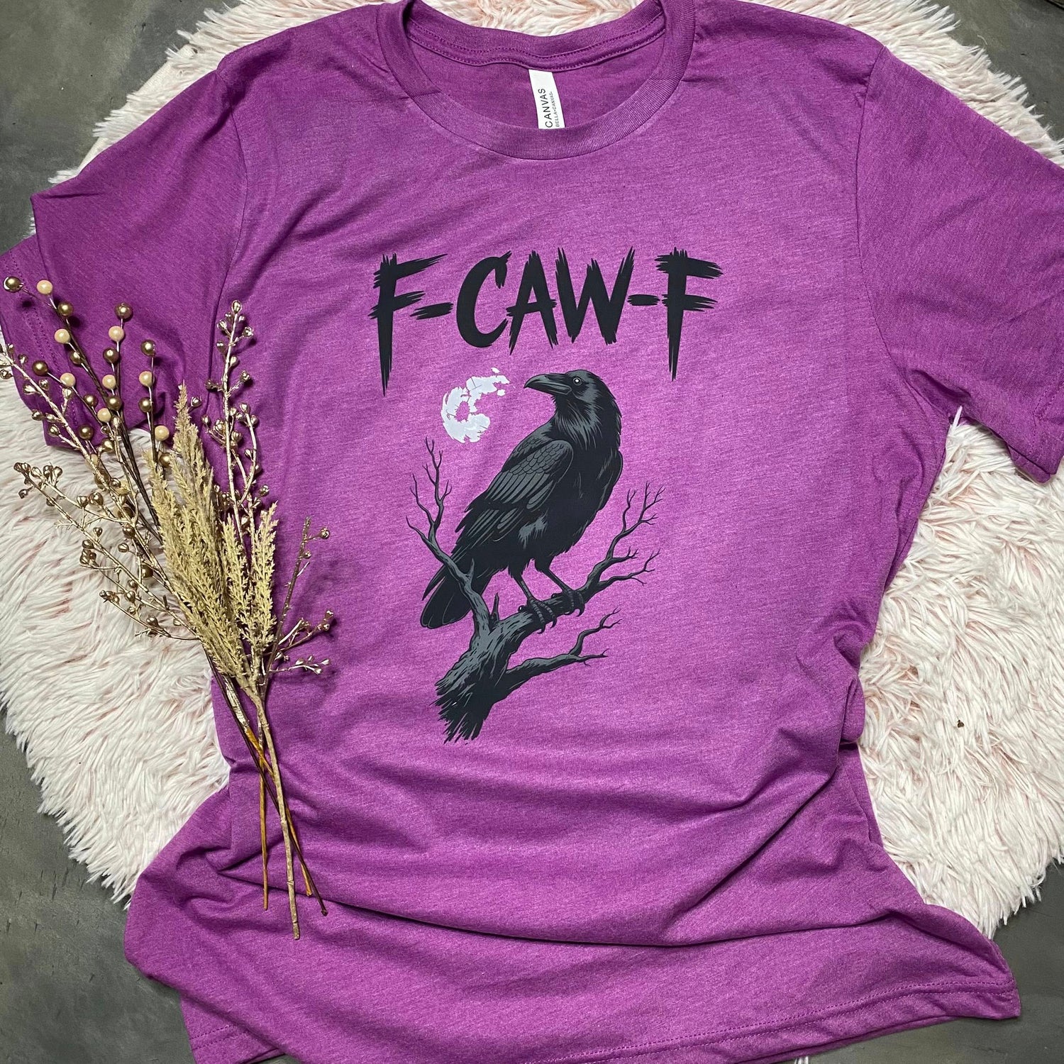 Purple t-shirt with a black crow graphic and 'F-CAW-F' text on a textured surface.