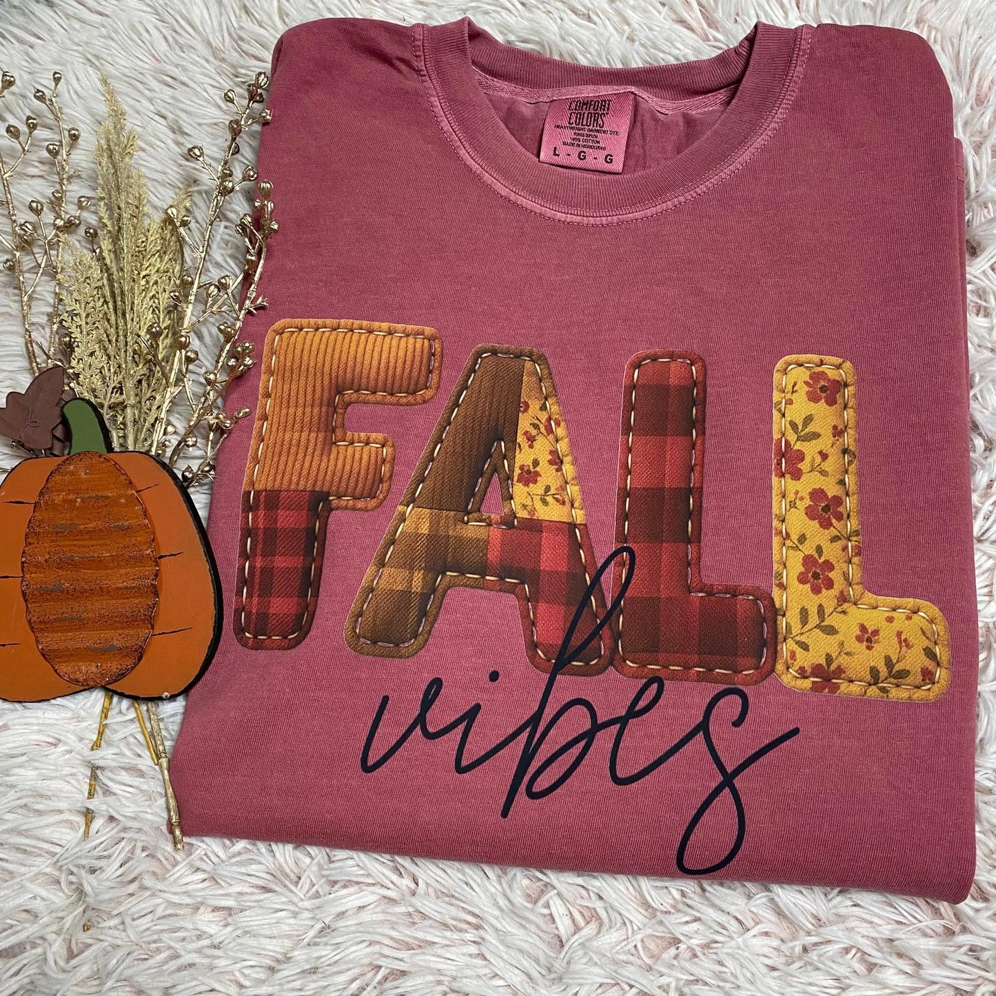 Folded maroon tee shirt with 'FALL vibes' text and patchwork design on a textured surface.