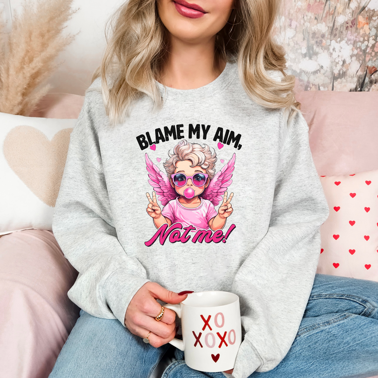 Cozy ash-colored sweatshirt featuring a trendy cupid illustration with pink wings, sunglasses, and bubblegum, captioned 'Blame My Aim, Not Me!' Perfect for adding a playful touch to your Valentine’s Day outfit or lounging in style.