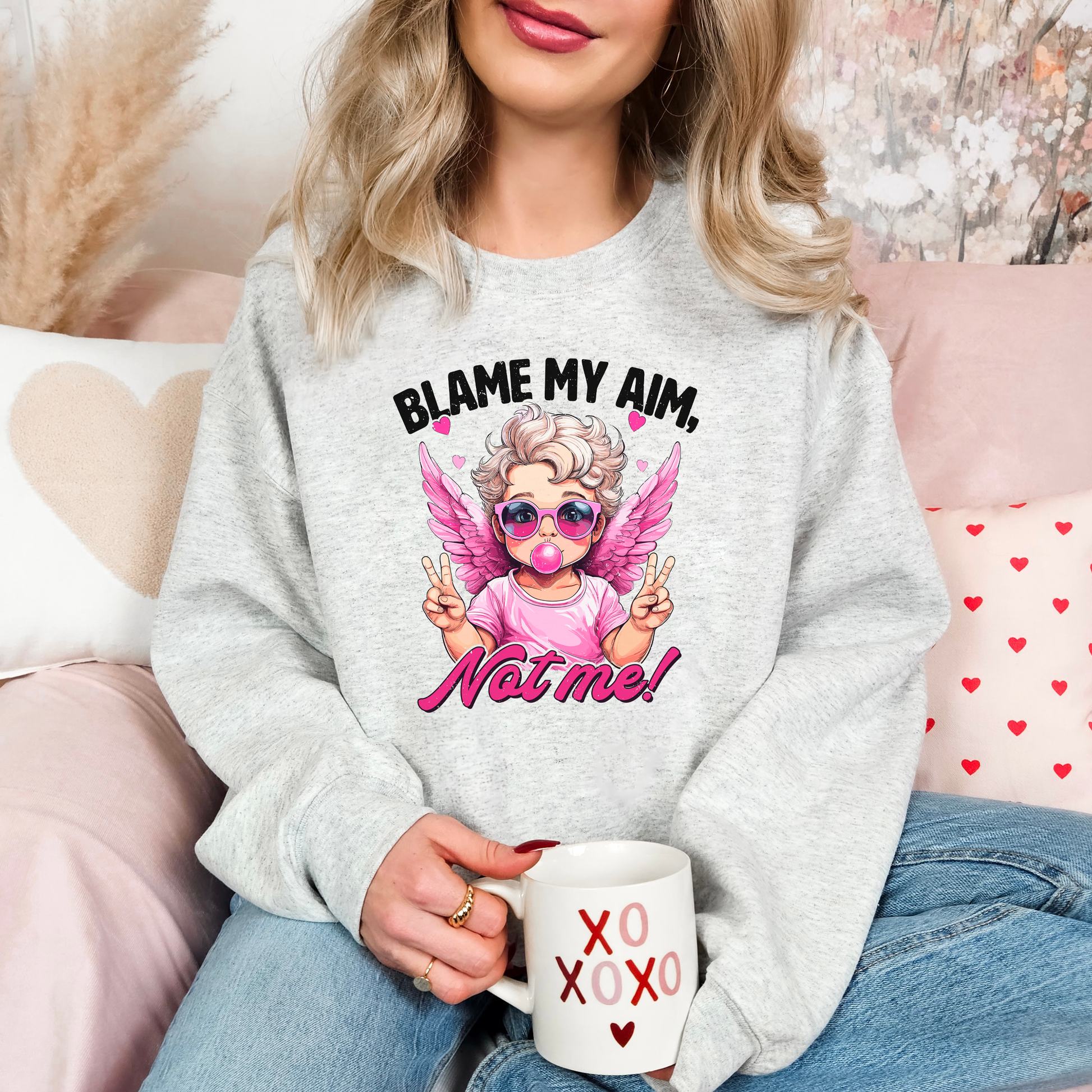 Cozy ash-colored sweatshirt featuring a trendy cupid illustration with pink wings, sunglasses, and bubblegum, captioned 'Blame My Aim, Not Me!' Perfect for adding a playful touch to your Valentine’s Day outfit or lounging in style.
