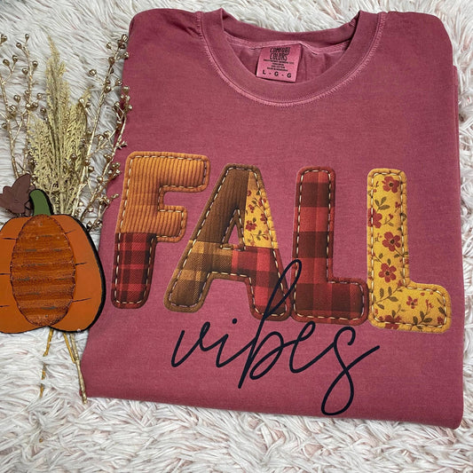 Folded maroon tee shirt with 'FALL vibes' text and patchwork design on a textured surface.