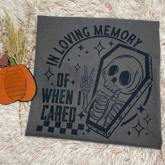 Gray graphic tee with skeleton design and text 'In loving memory of when I cared' on a textured surface.