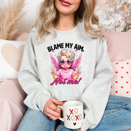Cozy ash-colored sweatshirt featuring a trendy cupid illustration with pink wings, sunglasses, and bubblegum, captioned 'Blame My Aim, Not Me!' Perfect for adding a playful touch to your Valentine’s Day outfit or lounging in style.