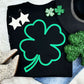 Black graphic tee featuring a large green puff outlined shamrock design, styled with glittery and festive St. Patrick’s Day accessories.
