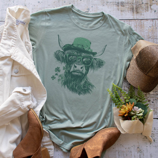 Dusty blue graphic tee featuring a Highland cow with a leprechaun hat, sunglasses, and clover, styled with rustic accessories.