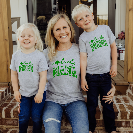 Heather gray matching St. Patrick’s Day graphic tees for mom and kids with 'Lucky Mama' and 'Lucky Mini' designs featuring green text and shamrocks.