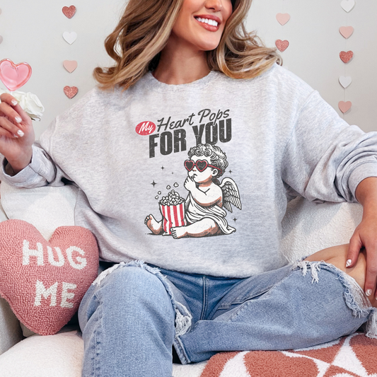 Comfortable ash-gray sweatshirt featuring a charming Cupid design holding a red and white striped popcorn bucket. The text reads 'My Heart Pops For You,' accented with playful typography and heart-shaped sunglasses, perfect for Valentine's Day.