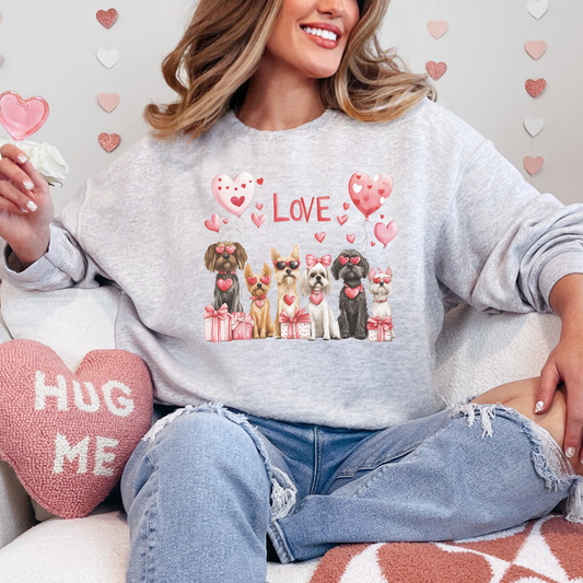 Adorable ash-gray sweatshirt perfect for Valentine's Day, featuring a group of cute dogs decorated with pink bows, heart-shaped sunglasses, and surrounded by gift boxes and heart-shaped balloons. The word 'LOVE' in bold pink font completes the festive look, making it an ideal outfit for dog lovers to celebrate the season of love.