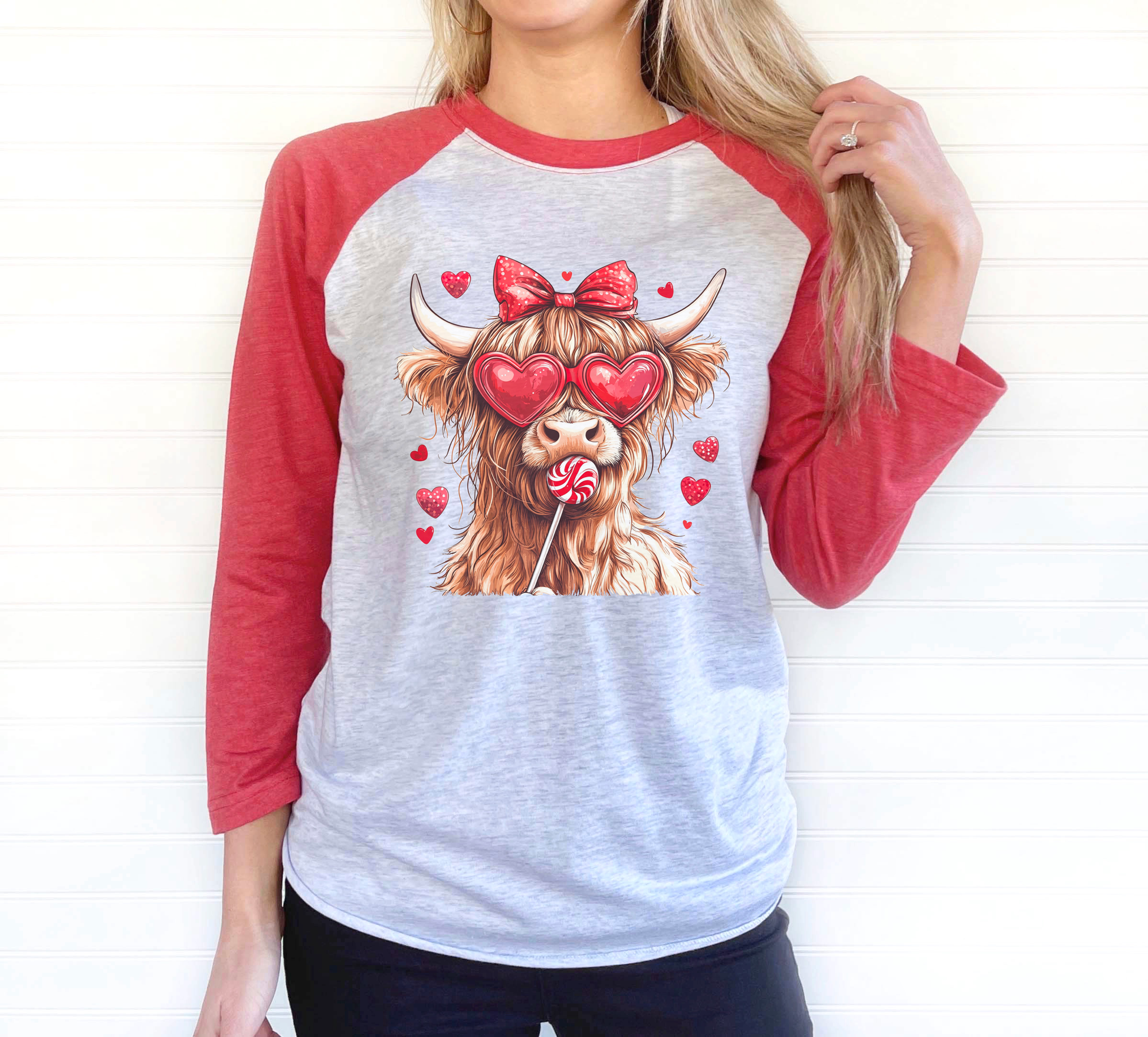 rendy raglan-style t-shirt with red sleeves and a gray torso, showcasing a playful highland cow design featuring heart-shaped sunglasses, a red bow, and a lollipop, surrounded by red hearts for Valentine's Day.